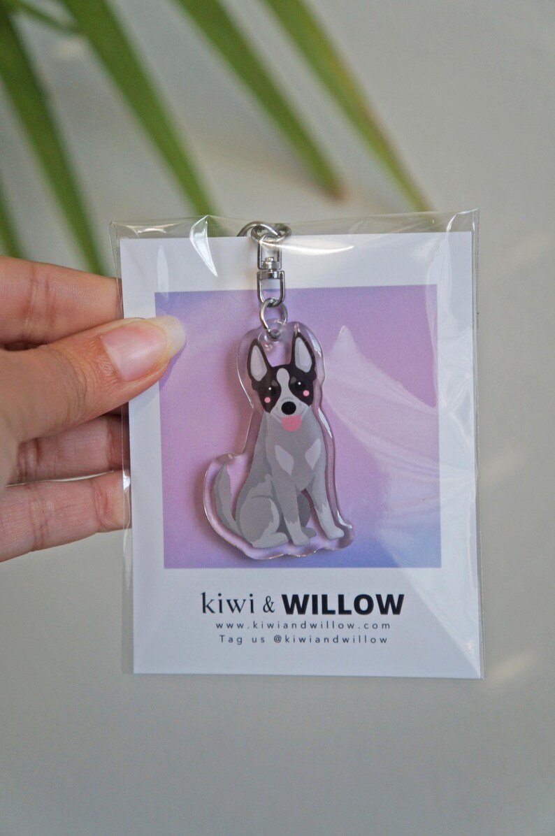 May include: A clear acrylic keychain featuring a cartoon dog with gray fur, black and white markings, and a pink tongue. The keychain is attached to a silver metal clasp. The packaging includes the text "kiwi & WILLOW" and a website address.