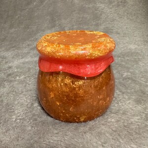 May include: A small, round jar with a textured, amber-colored body containing gold-toned flecks. The lid is a flat, circular piece in a shimmering orange-gold hue, featuring a decorative, draped, translucent red band around the upper rim.