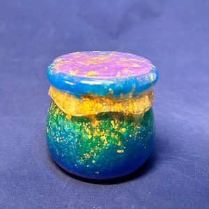 May include: A small, round resin jar with a lid, featuring a gradient of blue, green, and purple colors. The surface is embedded with gold leaf flakes throughout the body and lid, creating a shimmering, textured appearance with a glossy finish.