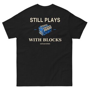 May include: A black short-sleeve t-shirt featuring a graphic of a blue engine block. The text reads STILL PLAYS WITH BLOCKS in cream lettering above and below the engine illustration, with the word GARAGEVERMIN printed in smaller text at the bottom.