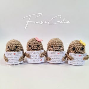 Può includere: Quattro figure di patata all'uncinetto, ognuna con un cartello con testo in italiano. Le patate hanno facce sorridenti e piccoli fiocchi rosa e gialli. I cartelli hanno le parole "PATATA POSITIVA".