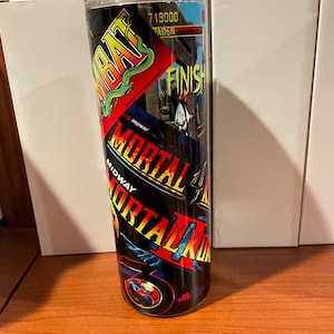 May include: A black and red tumbler with a Mortal Kombat theme. The tumbler features images from the video game, including the characters Scorpion, Sub-Zero, and Raiden. The text "Mortal Kombat" is printed on the tumbler in a bold, white font.