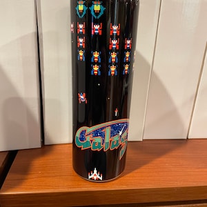 May include: A black stainless steel tumbler with a graphic of a spaceship and the text "Galaxian" on a black background. The graphic also includes pixelated spaceships and aliens in red, blue, and green.