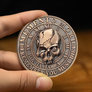 Op de afbeelding: Een koperkleurige munt met een schedel en gekruiste beenderen ontwerp. De munt heeft de woorden "Memento Mori" en "Remember You Must Die" gegraveerd rond de rand.