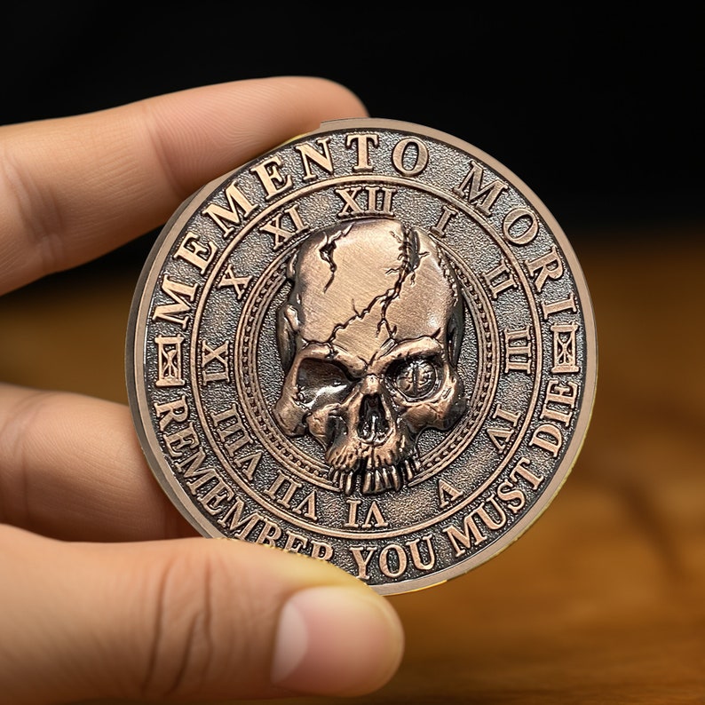 Op de afbeelding: Een koperkleurige munt met een schedel en gekruiste beenderen ontwerp. De munt heeft de woorden "Memento Mori" en "Remember You Must Die" gegraveerd rond de rand.