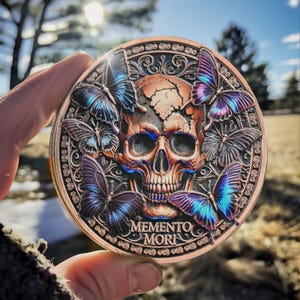 Op de afbeelding: Een ronde, koperkleurige munt met een gedetailleerd ontwerp met een schedel omringd door iriserende vlinders. De inscriptie "MEMENTO MORI" is zichtbaar aan de onderkant.