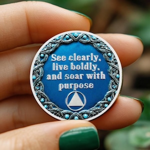 May include: A round, blue and silver coin with the text "See clearly, live boldly, and soar with purpose." The coin has a decorative border and a white triangle symbol.
