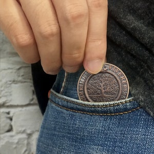 Op de afbeelding: Een koperkleurige munt met een levensboomontwerp en de woorden "Memento Mori Vivere" rond de rand. De munt wordt in een blauwe spijkerbroekzak gestopt.