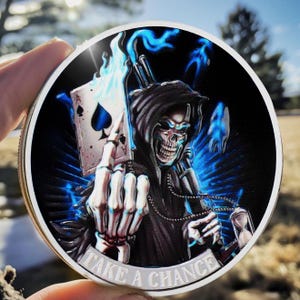 May include: A circular silver-coloured coin depicting a grim reaper holding an Ace of Spades card with blue flames. The coin has the text "TAKE A CHANCE" around the edge. The reaper has a skull face and wears a black hooded cloak.