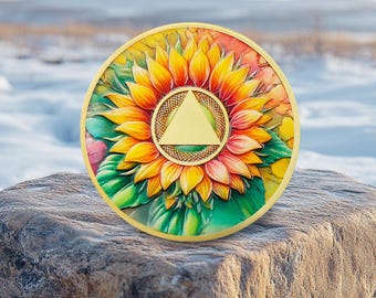 Könnte beinhalten: Ein kreisförmiges Medaillon mit einem leuchtend bunten Sonnenblumenmotiv mit gelben, orangefarbenen und grünen Blütenblättern. In der Mitte der Sonnenblume befindet sich ein glattes, goldfarbenes Dreieck in einem strukturierten, goldenen Rand.