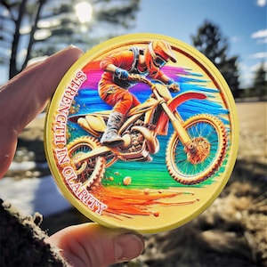 Può includere: Una moneta rotonda dorata con un'immagine colorata di un pilota di motocross su una moto da cross. Le parole "Strength in Clarity" sono scritte attorno al bordo della moneta.