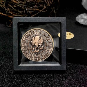 Op de afbeelding: Een bronzen munt met een schedelontwerp en de inscriptie "Memento Mori Remember You Must Die" rond de rand. De munt wordt getoond in een zwart vierkant frame met een transparante plastic hoes.