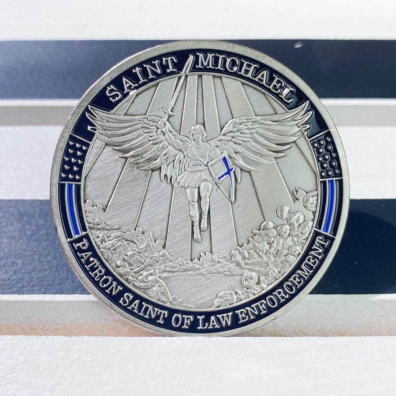 May include: Silver coin with a blue and black border. The coin features an image of Saint Michael, the patron saint of law enforcement, with a sword and shield. The text "Saint Michael Patron Saint of Law Enforcement" is inscribed around the edge of the coin.