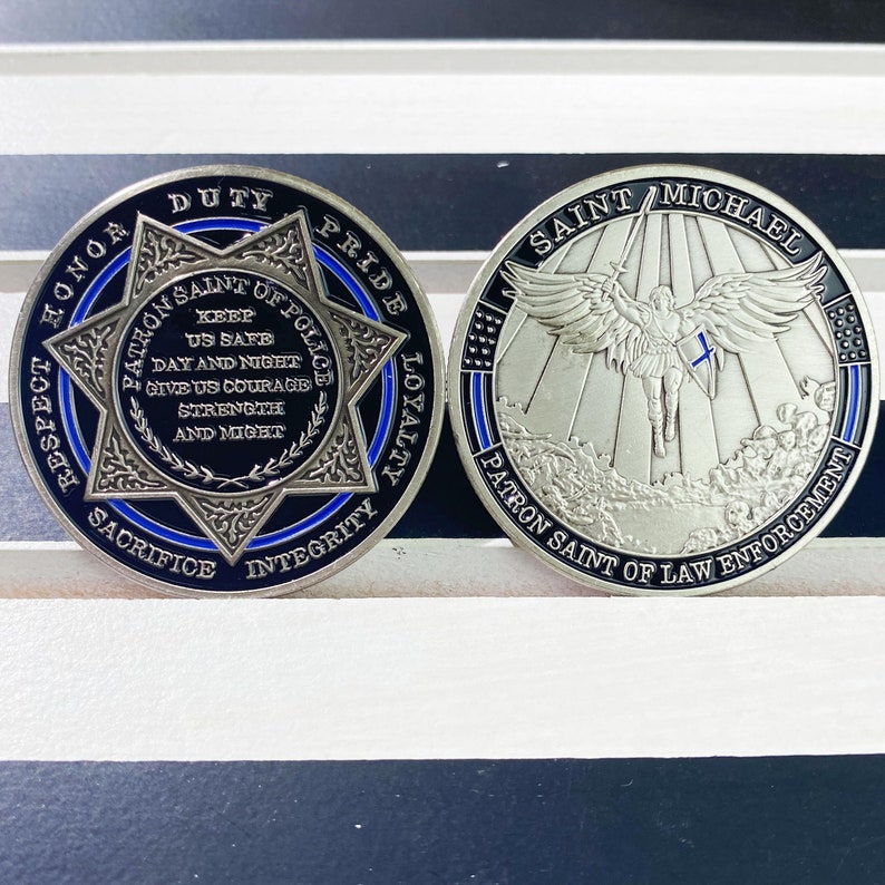 May include: A silver coin with a blue and black design featuring a star with the words "Patron Saint of Police" and "Keep Us Safe" around it. The other side of the coin features a silver image of Saint Michael with the words "Saint Michael Patron Saint of Law Enforcement".