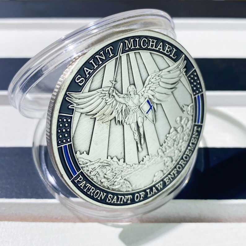 May include: Silver coin with a depiction of Saint Michael the Archangel with wings outstretched, holding a sword. The coin has the text "Saint Michael Patron Saint of Law Enforcement" around the edge.
