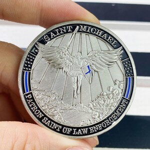 May include: Silver coin with a black border and blue accents. The coin features an image of Saint Michael, a winged angel, holding a sword and shield. The text "SAINT MICHAEL PATRON SAINT OF LAW ENFORCEMENT" is inscribed around the edge of the coin.