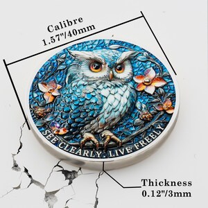 May include: A round, silver-colored coin with a detailed owl design. The owl is blue and white with orange eyes, surrounded by floral accents. The coin reads "SEE CLEARLY. LIVE FREELY." and has a diameter of 1.57 inches (40mm) and a thickness of 0.12 inches (3mm).