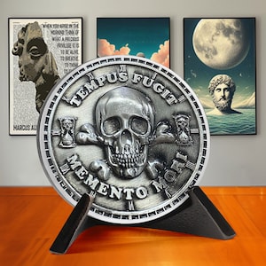 Op de afbeelding: Een zilverkleurige munt met een schedel en gekruiste beenderen ontwerp. De munt heeft de woorden "TEMPUS FUGIT" en "MEMENTO MORI" gegraveerd rond de rand. De munt staat op een zwarte standaard.