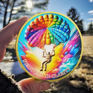 May include: A gold-colored round coin with a colorful parachute and a skydiver in the center. The coin has the words "Strength in Clarity" around the edge.