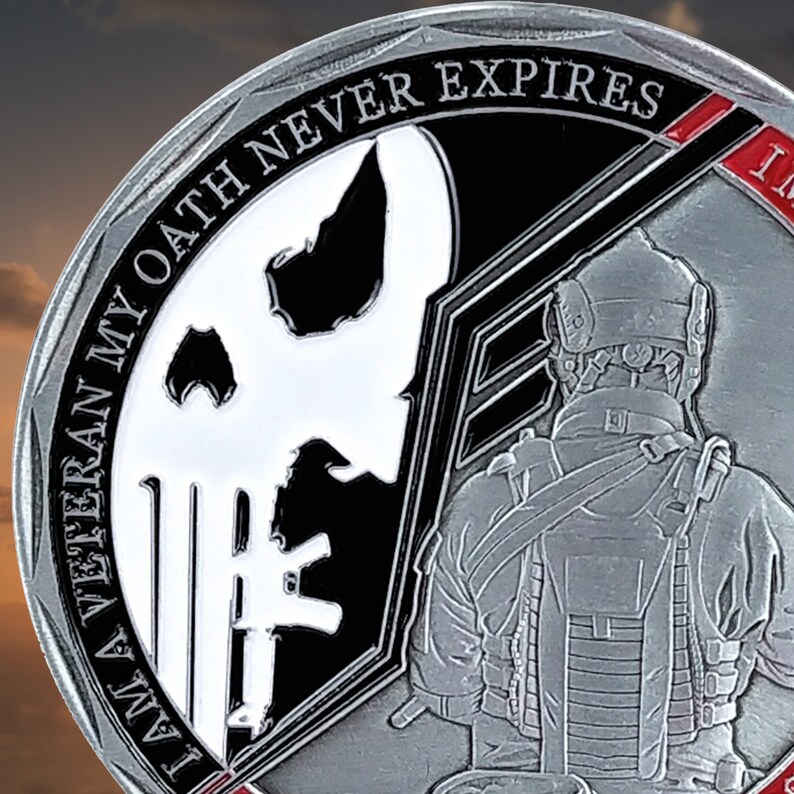 May include: A silver challenge coin with a black and white skull and crossbones design on one side and a soldier in tactical gear on the other. The coin has the text "I AM A VETERAN MY OATH NEVER EXPIRES" around the edge.