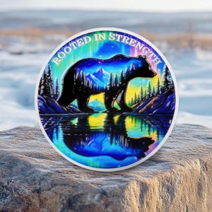 May include: A round, colorful graphic featuring a black bear silhouette against a landscape of mountains, trees, and a reflective lake. The text "ROOTED IN STRENGTH" arches around the top edge. The design has a vibrant, artistic style.