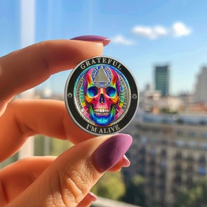 K&ouml;nnte beinhalten: Eine silberne M&uuml;nze mit einem schwarzen und regenbogenfarbenen Sch&auml;del-Design. Die M&uuml;nze tr&auml;gt die Worte "Grateful" und "I'm Alive". Der Sch&auml;del ist von einem Kreis mit den Worten "Unity, Service, Recovery" umgeben.