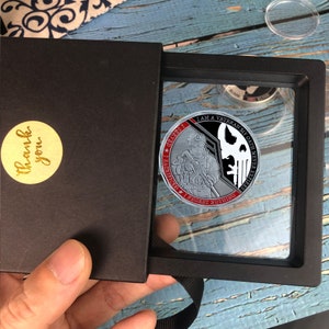 May include: A silver challenge coin with a black and white image of a soldier holding a rifle. The coin has red text that reads "I AM A VETERAN MY OATH NEVER EXPIRES.  I SACRIFICED I REGRET NOTHING.  GLORY I", and a skull with a crossbones.