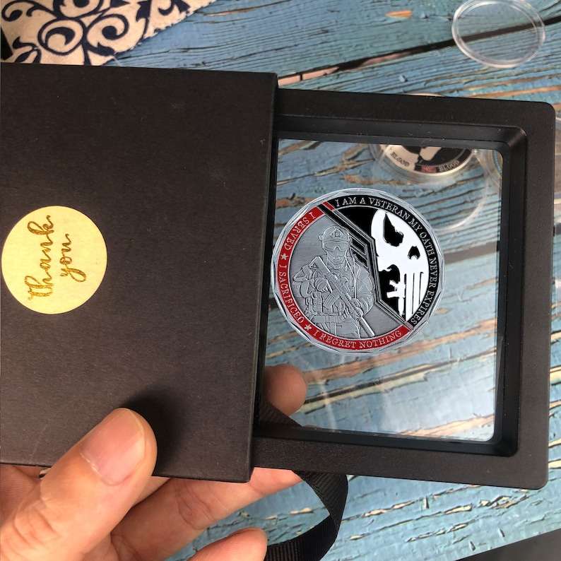 May include: A silver challenge coin with a black and white image of a soldier holding a rifle. The coin has red text that reads "I AM A VETERAN MY OATH NEVER EXPIRES.  I SACRIFICED I REGRET NOTHING.  GLORY I", and a skull with a crossbones.