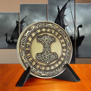 May include: A gold-coloured metal coin with a raised design of a hammer and runes. The coin is on a black stand. The coin has a circular shape and is surrounded by a decorative border.