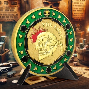 May include: A gold-coloured poker chip with a green and black border. The chip features a skull with a red mohawk and a gun pointed at its head. The text "YOU LOSE!" is written around the skull. The chip is on a black stand.