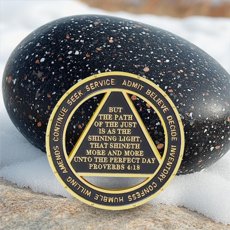 Puede incluir: Una moneda negra y dorada con el texto "BUT THE PATH OF THE JUST IS AS THE SHINING LIGHT, THAT SHINETH MORE AND MORE UNTO THE PERFECT DAY PROVERBS 4:18". La moneda est&aacute; colocada frente a una piedra negra moteada.