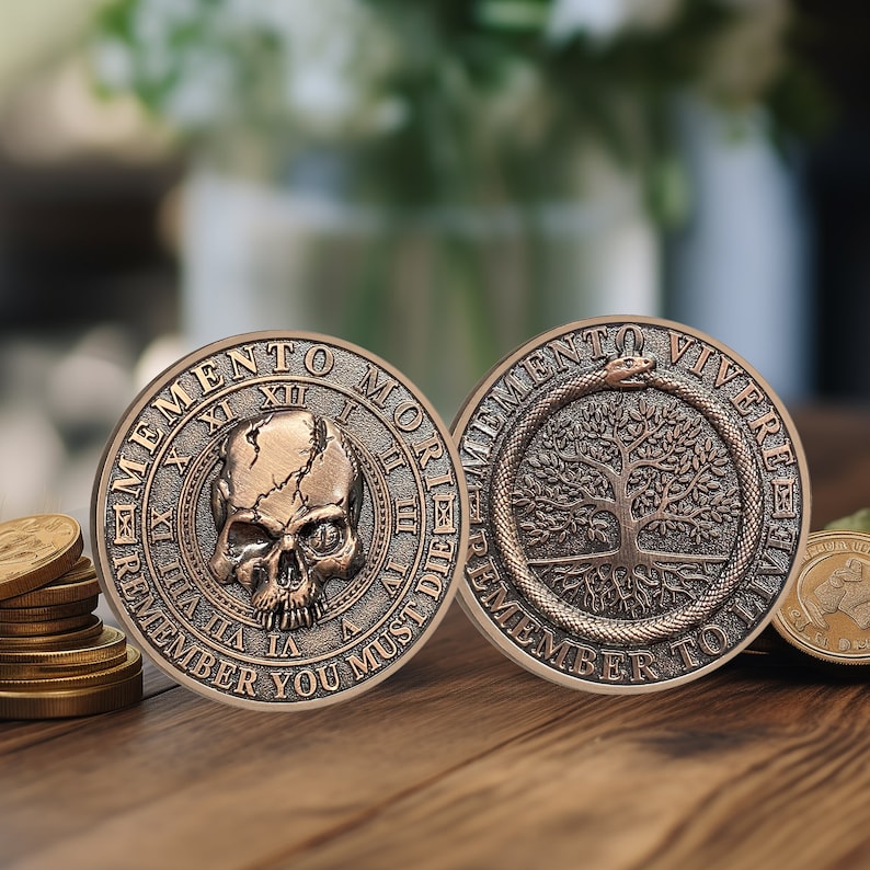Op de afbeelding: Twee ronde, koperkleurige munten met verhoogde randen. De eerste munt heeft een schedel met een gebarsten gezicht en de woorden "Memento Mori" en "Denk eraan dat je moet sterven". De tweede munt heeft een boom met wortels en een slang die om de stam is gewikkeld met de woorden "Memento Vivere" en "Denk eraan om te leven".
