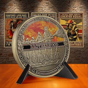 May include: A round, silver-coloured coin with a black and red design. The coin features a group of firefighters in action, with the words "VALOR", "SERVICE", "HONOR", "COURAGE", "BROTHERHOOD", and "DEDICATION" inscribed around the edge. The coin is displayed on a black stand.