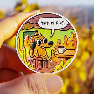 May include: A circular enamel pin featuring a cartoon dog wearing a hat, sitting at a table with a coffee mug while surrounded by orange and yellow flames. A white speech bubble above the dog contains the black text, THIS IS FINE.