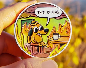 May include: A circular enamel pin featuring a cartoon dog wearing a hat, sitting at a table with a coffee mug while surrounded by orange and yellow flames. A white speech bubble above the dog contains the black text, THIS IS FINE.