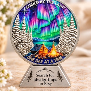 Könnte beinhalten: Ein kreisförmiges Metalldisplay mit einer farbenfrohen Nordlichtszene, Tannenbäumen, einem Zelt und einem Lagerfeuer. Der Text lautet GUIDED BY THE LIGHT und ONE DAY AT A TIME. Der Sockel enthält den Text Search for idealgiftings on Etsy.