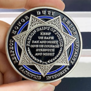 May include: A silver and black police challenge coin with a blue line around the edge. The coin features a six-pointed star with the words "Patron Saint of Police" and "Keep Us Safe Day and Night Give Us Courage Strength and Might" inscribed inside. The words "Respect Honor Duty Pride Loyalty Sacrifice Integrity" are inscribed around the edge of the coin.