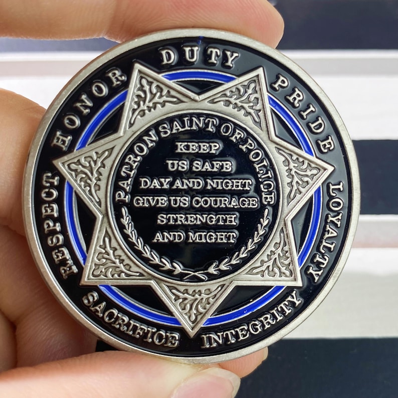 May include: A silver and black police challenge coin with a blue line around the edge. The coin features a six-pointed star with the words "Patron Saint of Police" and "Keep Us Safe Day and Night Give Us Courage Strength and Might" inscribed inside. The words "Respect Honor Duty Pride Loyalty Sacrifice Integrity" are inscribed around the edge of the coin.