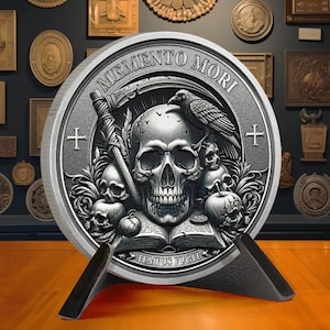 Op de afbeelding: Een zilveren munt met een schedel en kruisbeenderen ontwerp. De munt heeft de woorden "Memento Mori" en "Tempus Fugit" rond de rand. De munt staat op een zwarte standaard.