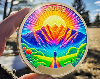 Può includere: Una moneta rotonda dorata con un'immagine colorata di una catena montuosa e una persona che cammina verso un'alba. La parola "SOBER" è stampata in lettere dorate nella parte superiore della moneta.