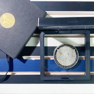 May include: A silver coin with a blue and white ribbon design. The coin features an image of Saint Michael, the patron saint of law enforcement, with wings spread and a sword in his hand. The text "Saint Michael Patron Saint of Law Enforcement" is inscribed around the edge of the coin.