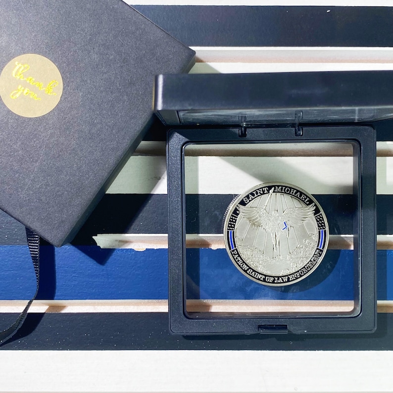 May include: A silver coin with a blue and white ribbon design. The coin features an image of Saint Michael, the patron saint of law enforcement, with wings spread and a sword in his hand. The text "Saint Michael Patron Saint of Law Enforcement" is inscribed around the edge of the coin.