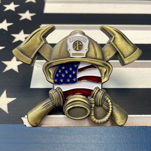 Puede incluir: Una moneda de desafío de bombero de metal dorado que presenta un casco de bombero con un diseño de bandera estadounidense, hachas cruzadas y una máscara de gas.