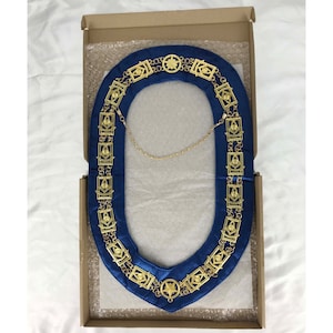 Puede incluir: Un collar masónico dorado con respaldo de terciopelo azul. El collar presenta un diseño de estrella y cuadrado.