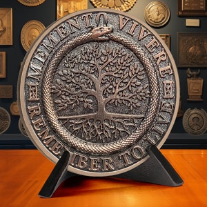 Op de afbeelding: Een ronde bronzen plaquette met een levensboomontwerp en een slang eromheen gewikkeld. De plaquette heeft de woorden "Memento Vivere Remember To Live" rond de rand gegraveerd. De plaquette staat op een zwarte standaard.