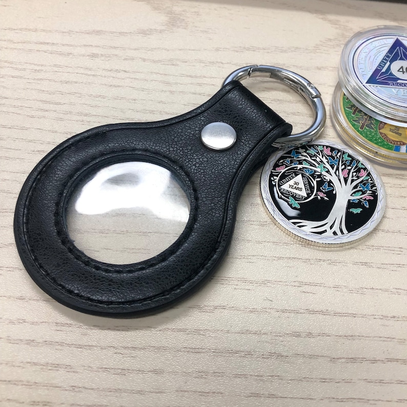 K&ouml;nnte beinhalten: Schwarzer Leder-M&uuml;nzhalter mit einem durchsichtigen Fenster und einem silbernen Ring. Der Halter ist an einem silbernen Schl&uuml;sselring befestigt. Eine silberne M&uuml;nze mit einem Baum und Schmetterlingen ist im Hintergrund sichtbar.