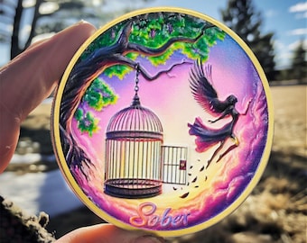 May include: A round, gold-rimmed coin with a colorful image of a bird flying out of a cage. The background is a vibrant purple and pink sunset with a green tree in the upper left corner. The word "Sober" is written in gold at the bottom of the coin.