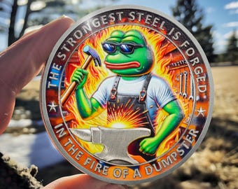May include: A silver round coin with a green cartoon frog wearing sunglasses and holding a hammer. The frog is standing in front of a glowing orange anvil. The coin has the text "The strongest steel is forged in the fire of a dumpster" around the edge.