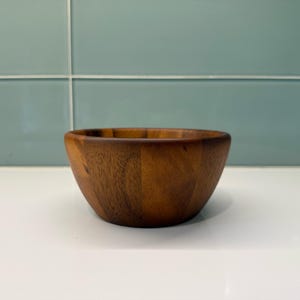 Peut inclure: Un petit bol rond en bois de couleur marron foncé. Le bol est fabriqué à partir d'une seule pièce de bois et présente une finition lisse et polie. Il repose sur une surface blanche, devant un fond de carreaux bleu clair.
