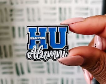 May include: A rectangular pin featuring the letters "HU" in blue and white, with "Hampton" and "Alumni" in white text. The pin has a silver border. The pin is held up by a person.